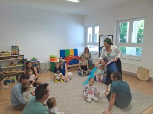 A képen a zeneterápiás foglalkozás indulása látható, a gyerekek a szüleikkel egy körben ülnek a szőnyegen, miközben a terapeuta kiosztja a színes kendőket a foglalkozáshoz.