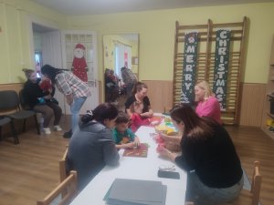 A képen a gyerek és pedagógusaink egy asztalnál ülve készítik, só-liszt gyurmából a karácsonyi formákat.