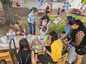 A képen a Mezőtúri Tagintézményben megrendezésre került Víz-Party közös kézműves foglalkozása látható a tagintézmény udvarán. A gyerekek, szüleik és pedagógusaink segítségével egy asztalt körbeülve kézműveskednek.