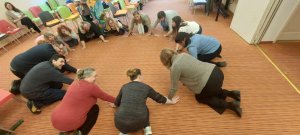 A képen egy figyelemkoncentrációt fejlesztő mozgásos gyakorlat saját élményű kipróbálása látható. A résztvevők és az előadók a földön guggolva végzik a gyakorlatokat közösen egy kört alkotva.