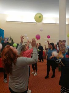 A képen egy figyelemkoncentrációs gyakorlat végzése látható léggömbök segítségével. A résztvevők a teremben, párokat alkotva ütögetik egymásnak a színes léggömböket.