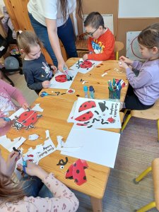 A képen a gyermekek asztal körül ülnek. A katica sablonját kivágják, majd a logopédus segítségével a megfelelő helyre ragasztják. A végére reptethető katicák készülnek el.