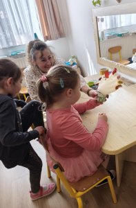 Az asztalnál ülve egy kislány az elkészült tollakkal díszített gumikesztyű tyúkanyót fújja fel a pohárba szúrt szívószállal. Másik kislány kedvet kap a tevékenységhez és éppen leül az asztalhoz.