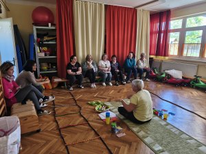 A képen az aktív poharak a gyakorlatban bemutatása látható. A szakember a terem közepén egy szőnyegen ülve tartja az előadást a vele szemben egy tornapadon helyet foglaló hallgatóságnak. 