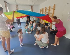 A képen a szülők körben egy szivárványszínű óriáskendőt fognak, melyet fel-le emelgetnek. A gyerekek a kendő alatt csücsülnek, szaladnak át.