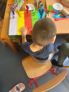 A képen egy gyermek látható a kis asztalnál ülve, előtte egy kartondoboz aminek a teteje öt különböző színű, egyenlő méretű sávra van osztva. (fehér, citromsárga, piros, zöld és kék). A sávokon megtalálható helyekre kell a gyermeknek a megfelelő színű színes virágot elhelyeznie.  