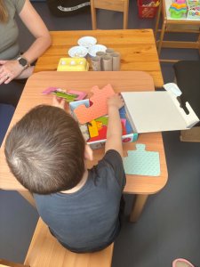 A képen egy gyermek látható a kis asztalnál helyet foglalva amint válogatja a színes puzzle darabokat az előtte lévő dobozból.