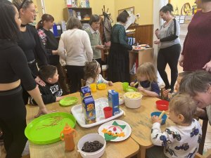 A képen a gyermekek egy asztalt körbeülve fogyasztják az uzsonnát, amíg a körülöttük lévő szülők beszélgetnek. 