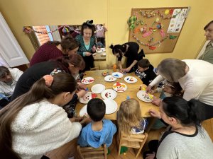 A képen a gyerekek és szüleik közösen készítik papírtányérokból a színes bohócokat egy asztalt körbeülve.