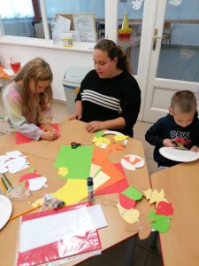 Egy anyuka és két kisgyerek dolgozik az asztalnál ülve. A kislány nyírja a faleveleket a sablonok alapján, a kisfiú a papírtányérból a keretet vágja. A kép előterében a mintadarab látható.