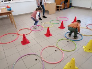 A képen a két csapatból egy-egy gyermek szökdel a színes karikákba miközben a karikák mellett levő gesztenyéket berakják a karikákba. Egymás mellett haladnak. A képen a két karika sor között látható piros bójákat kerülgetve kell visszafutniuk a csapatukhoz.