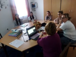 A logopédus háttal látszik a képen, amint a laptop és kivetítő segítségével magyaráz. Az asztalon láthatók fejlesztő eszközök. 5 óvodapedagógus figyeli az előadást.