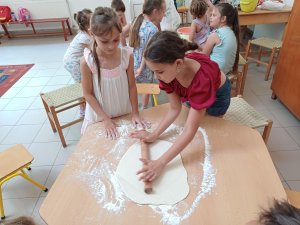 A képen a só-lisztgyurmát egy nagylány nyújtja sodrófával az asztalon. A háttérben a másik asztalnál szaggatják a korongokat.