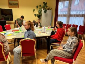 A képen a foglalkozáson résztvevő gyermekek láthatóak amint az előadó teremben az asztal körül különböző feladatokat végeznek.