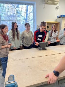 A képen az egyik tanuló megpróbál felemelni egy asztalról egy üllőt. Körülötte öten figyelik.