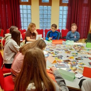 A képen a tanulók és a foglalkoztatást tartó pedagógus látható, egy asztal körül. Előttük az asztalon színes képek láthatóak szétterítve. 