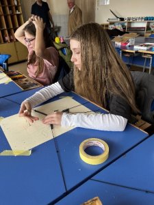 A képen két tanuló készíti rajz műveit egy-egy asztalnál ülve.