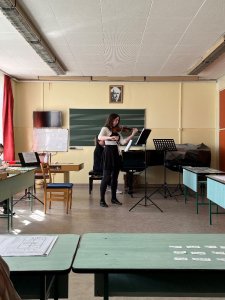 A képen az egyik tanuló látható, egy osztályteremben. A tábla előtt állva hegedűjén játszik. 