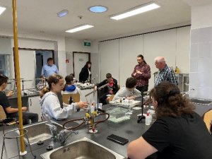 A képen a tanulók egy kémia laborban hallgatják az előadót és közben az utasításai szerint érdekes kísérleteket folytatnak. 