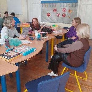 A képen a pedagógusok láthatóak egy asztalnál ülve amíg a gyerekek a háttérben egy másik asztalnál végzik a kiscsoportoknak kiosztott feladatokat.