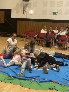 A képen a gyermekek és az előadó végzik közösen a gyakorlatot a kék szőnyegen.