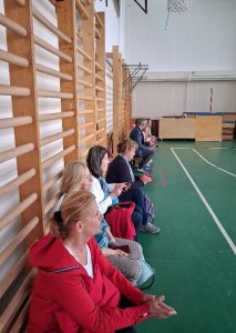 A képen a tornateremben, a bordásfalak előtt helyet foglaló résztvevők egy része látható amint figyelik a bemutató órát.