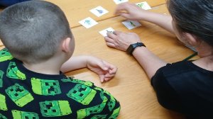 A képen egy gyermek és egy pedagógus látható az asztalnál, háttal a fényképésznek, miközben a logopédiai fejlesztő kártyajátékot használják. 