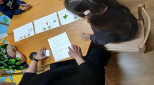 A képen egy asztal körül három gyermek és egy pedagógus áll. előttük három papír látható (az első papíron 1 db méhecske, a második papíron 2 db méhecske és a harmadik papíron 3 db méhecske) ezekre a papírokra kerültek a szó kártyák a szótagszám alapján.