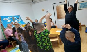 A képen a gyerekek mozgással összekötött mondókás gyakorlatot végeznek közösen a pedagógussal.