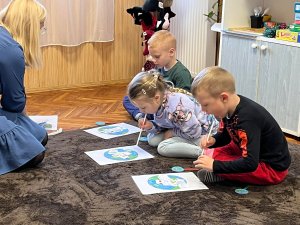 A képen három gyermek szívószálaik segítségével fújógyakorlatokat végeznek törökülésben egy szőnyegen.