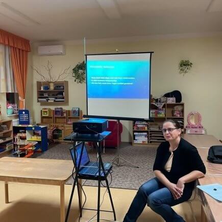 A képen „A mese hatása a gyermek lelki egészségére” című előadás pillanatai láthatóak egy tanteremben, a projektor mellett ülő előadó éppen az előadását tartja, háttérben a kivetített prezentáció.