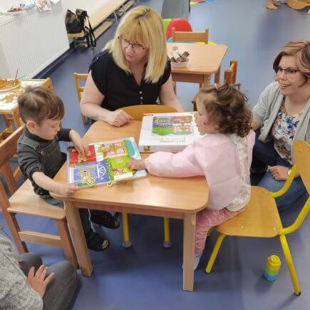 A képen két gyermek és két felnőtt látható, amint egy kis asztalt körbeülve Finommotorika és szem-kéz koordináció fejlesztése puzzlet használnak.