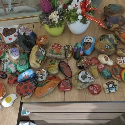 A képen a gyermekek által festett sok sok színes kavics látható egy asztalon. A kavicsokon látható tulipán, katicabogár, szívecske, sündisznó, bagoly, gomba, cica, polip és kisegér is.