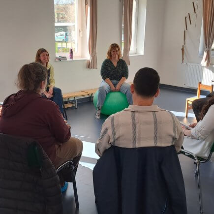 A kép a szülői értekezlet egyik pillanatát örökíti meg, amint a résztvevők egy nagy körben ülnek és az egyik édesanya éppen megosztja gondolatait, aggodalmait a többiekkel.