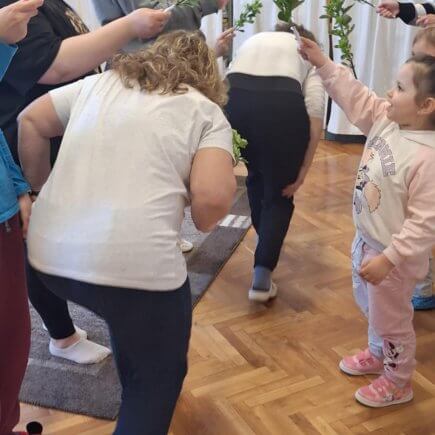 A képen "Bújj, bújj zöld ág" játékot játszanak a szülők és a gyermekek közösen. Az "alagutat" a gyermekek és a szülők közösen alakították ki egy-egy zöld ágacska segítségével, amit a magasba tartottak.