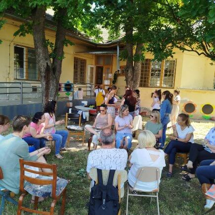 A kép a Mezőtúri Tagintézményben megrendezett szakmai beszélgetés pillanatát örökíti meg, amint a munkatársak és vendégeik egy nagy körben ülnek kint a szabadban a fák árnyékában.