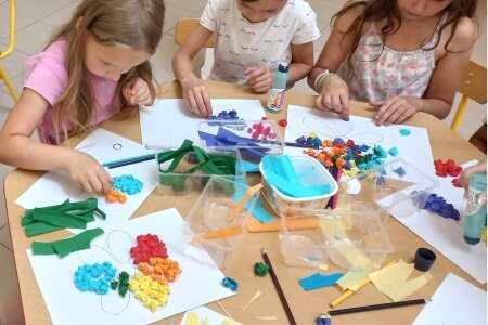 A képen a kislányok az asztalnál ülnek, és szorgosan sodorgatják a krepp-papír golyókat, amiket felragasztanak a megrajzolt virágra. Élénk színű szirmok készülnek a Hétszínvirág című mese illusztrációjaként.