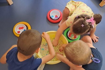 A képen a kincskeresés pillanatai láthatóak amint három kisgyermek keresi a szívecskéket a hidrogolyók között.