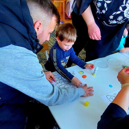 A képen a gyermek egy asztalnál állva szülői segítséggel színes, tavaszi pillangót készít ragasztással.