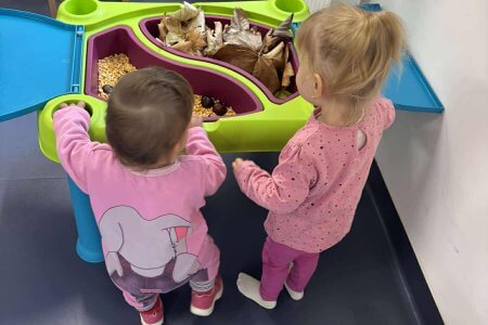 A képen két gyermek látható akik egy asztal mellett állva nézegetik a kihelyezett őszi terméseket.