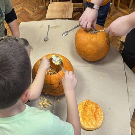 A képen a gyerekek készítik a töklámpásaikat pedagógusunk segítségével egy asztal körül, éppen a tökmagokat szedik ki egy kanállal.
