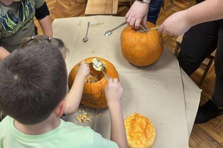 A képen a gyerekek készítik a töklámpásaikat pedagógusunk segítségével egy asztal körül, éppen a tökmagokat szedik ki egy kanállal.