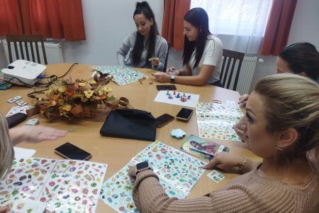 Az asztalnál ülve kipróbálják az óvónők a fejlesztő játékot. A lapokon látható sok kép közül a húzott kártyán levő 3 képet kell megkeresni. A felnőttek számára is élvezetes a saját készítésű játék.