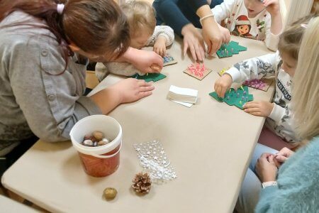 A képen a gyermekek karton karácsonyfákat díszítenek csoportosan egy asztalt körbe ülve.