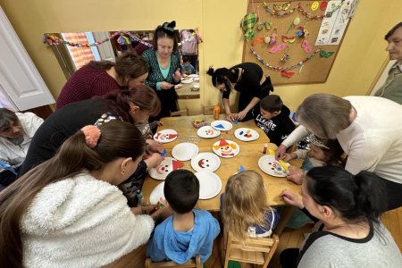 A képen a gyerekek és szüleik közösen készítik papírtányérokból a színes bohócokat egy asztalt körbeülve.