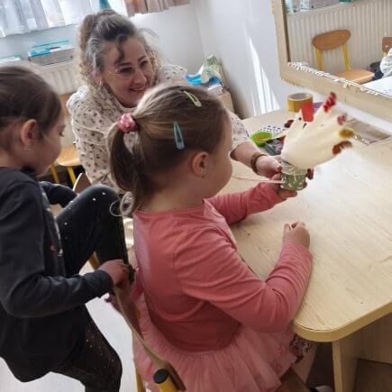 Az asztalnál ülve egy kislány az elkészült tollakkal díszített gumikesztyű tyúkanyót fújja fel a pohárba szúrt szívószállal. Másik kislány kedvet kap a tevékenységhez és éppen leül az asztalhoz.