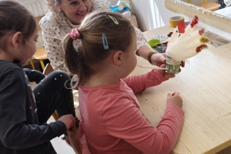 Az asztalnál ülve egy kislány az elkészült tollakkal díszített gumikesztyű tyúkanyót fújja fel a pohárba szúrt szívószállal. Másik kislány kedvet kap a tevékenységhez és éppen leül az asztalhoz.