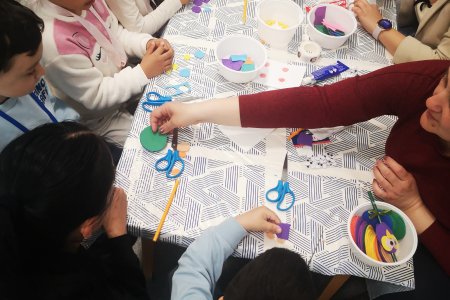 A képen a gyerekek egy asztalt körbeülve, színes formákat vágnak, amelyekből a foglalkozás végére összeáll a saját kis alkotásuk.