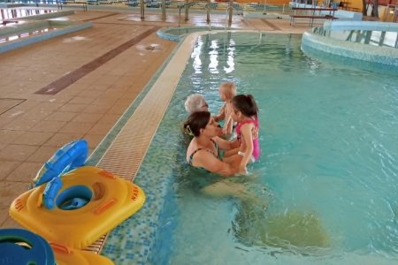 A kép az uszodában készült amint két szülő a medence szélén ülve a gyerekekkel az ölükben, a játékos átmozgató foglalkozáson vesznek részt.