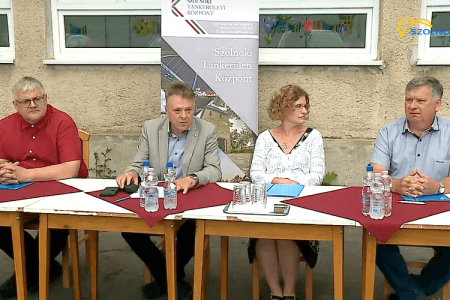 A kép az RRF-1.2.4-25-2025-00049 kódszámú, „Kiemelt figyelmet igénylő gyermekek és tanulók minőségi ellátása a Szolnoki Tankerületi Központban” című pályázat keretében beszerzett két, Hyundai Kona típusú, teljesen elektromos meghajtású gépjármű átadásán készült, és az átvevők láthatóak rajta egy asztalnál ülve.